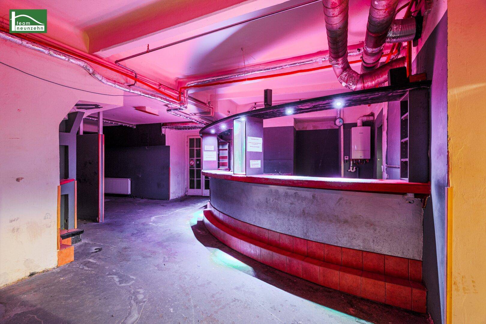 Bar im Untergeschoss mit geschwungener Theke, roten Akzenten und sichtbaren Lüftungsrohren unter pinker Beleuchtung.