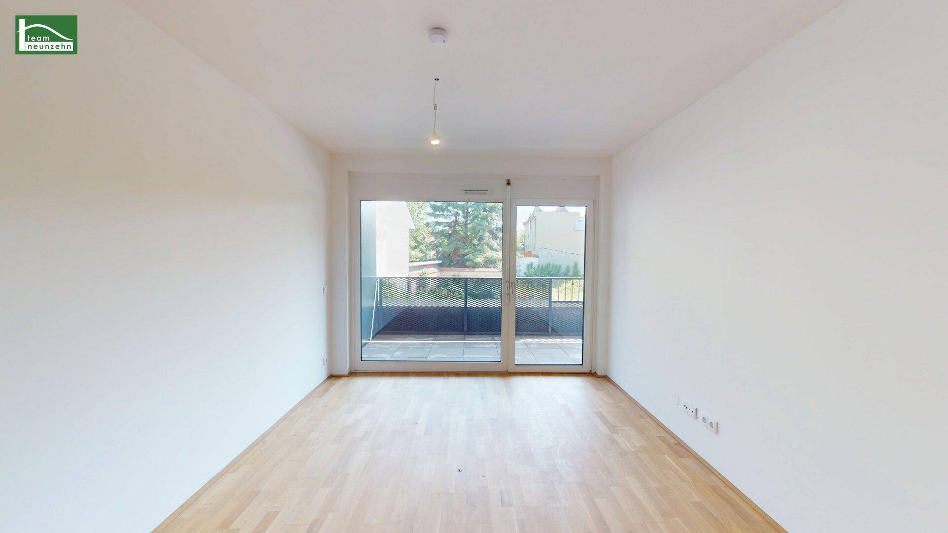 Leerer Wohnraum mit Holzboden und großen Fenstertüren, die auf einen Balkon führen und viel Licht hereinlassen.