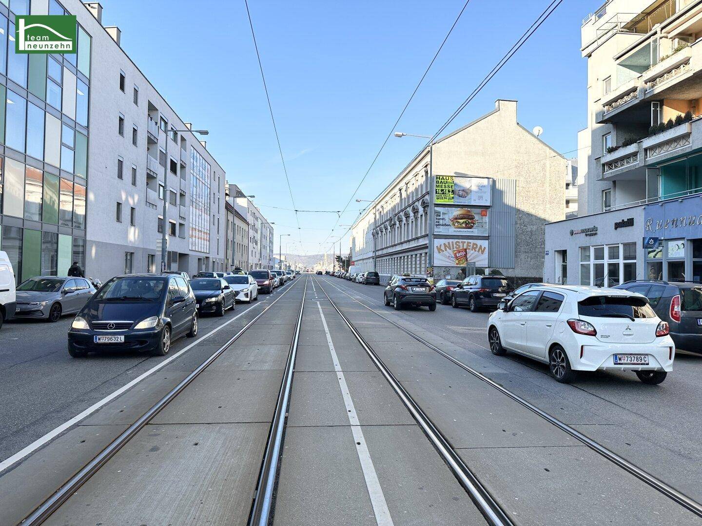 Belebte Stadtstraße mit Straßenbahnschienen, parkenden Autos und mehrstöckigen Gebäuden unter blauem Himmel.