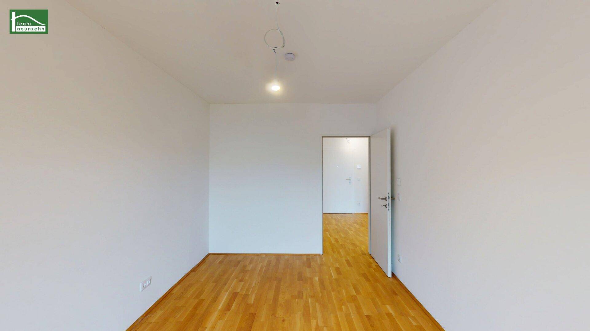 Leerer Raum mit hellem Parkettboden und offener Tür zu einem weiteren Zimmer.