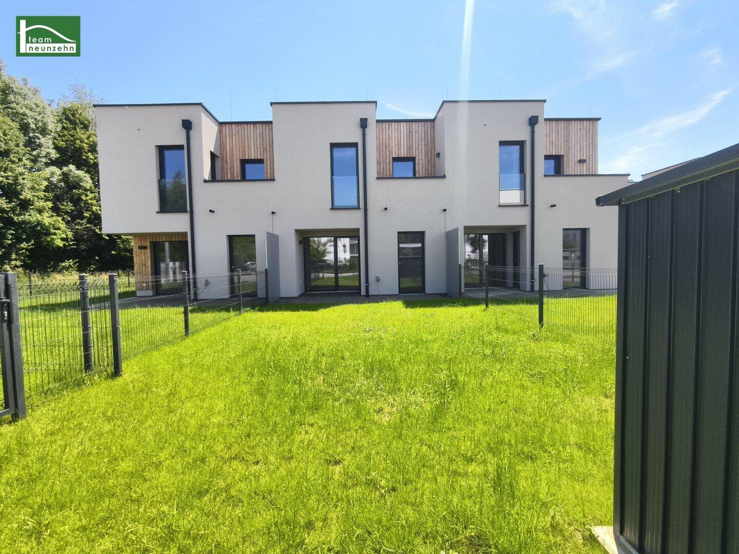 Moderne Reihenhäuser mit heller Fassade, Holzdetails und einem grünen Gartenbereich unter blauem Himmel.
