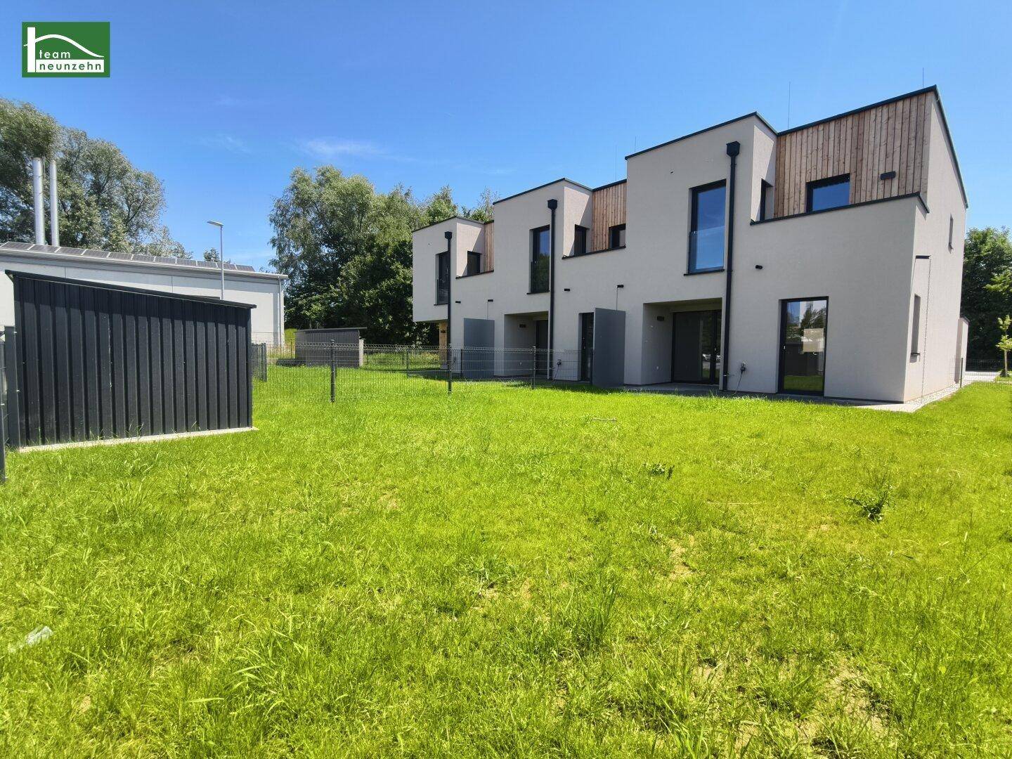 Moderne Reihenhäuser mit heller Fassade und Holzverkleidung, umgeben von einer weitläufigen Rasenfläche unter blauem Himmel.