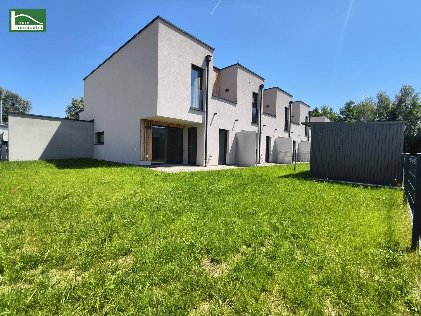 Moderne Reihenhäuser mit heller Fassade, großen Fenstern und einem weitläufigen Rasenbereich unter blauem Himmel.