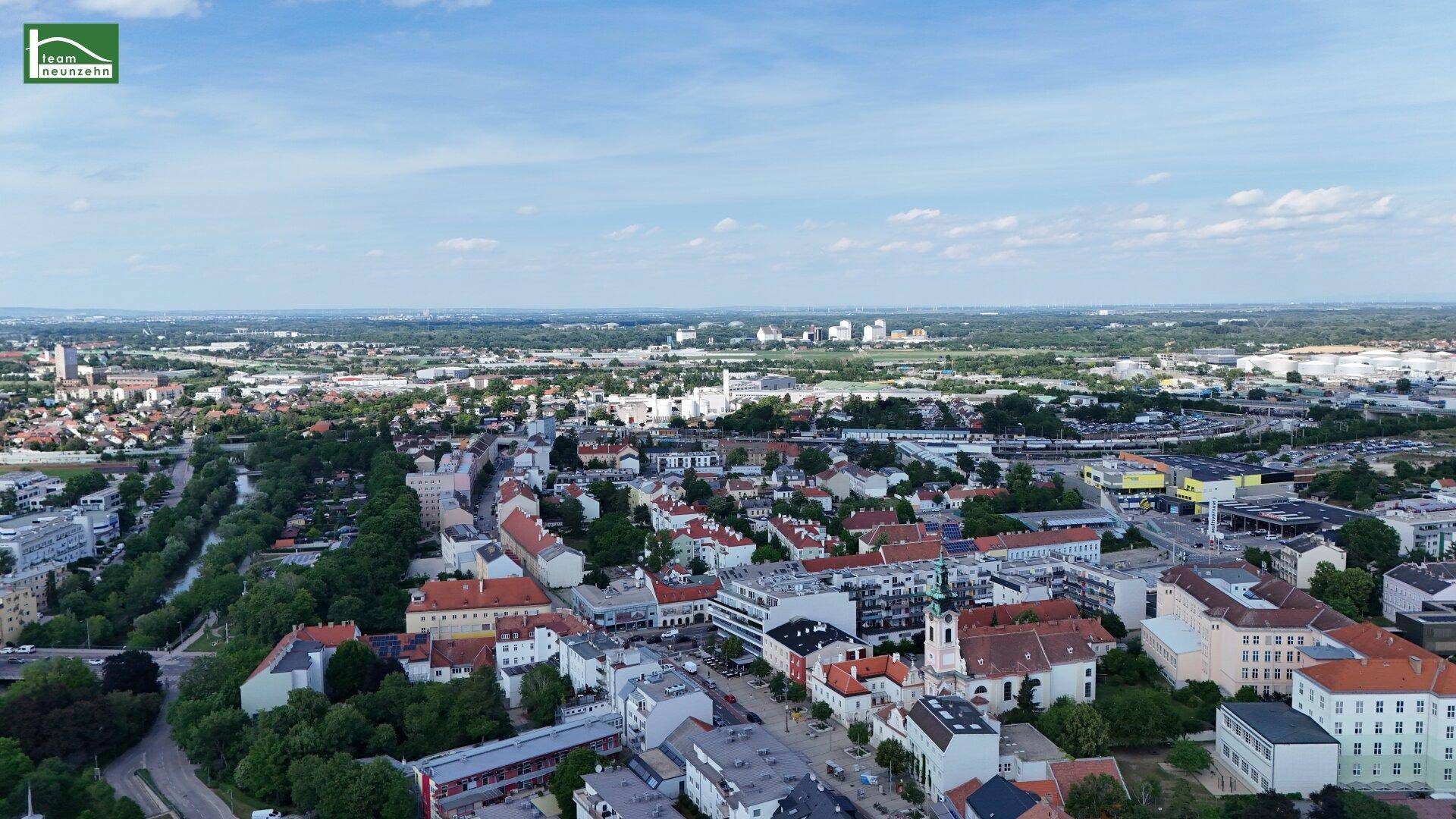 Luftaufnahme einer Stadt mit Wohngebäuden, Grünflächen und einem Fluss unter blauem Himmel.