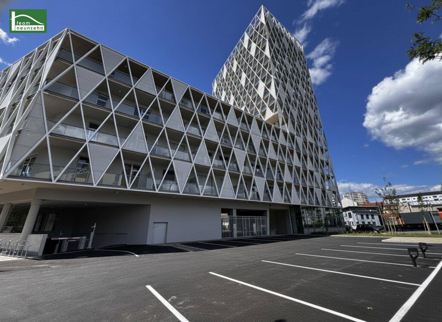 Modernes Gebäude mit auffälliger geometrischer Fassade und einem davorliegenden Parkplatz unter blauem Himmel.