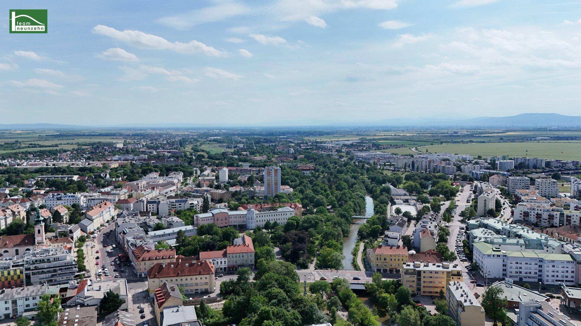 Luftaufnahme einer Stadt mit Wohngebäuden, viel Grün und einem Fluss, umgeben von ländlicher Landschaft.