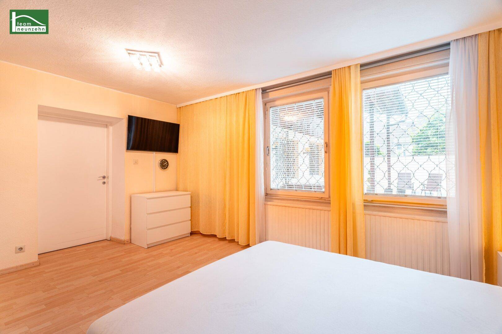 Schlafzimmer mit hellem Parkettboden, gelben Vorhängen und einem an der Wand montierten Fernseher.