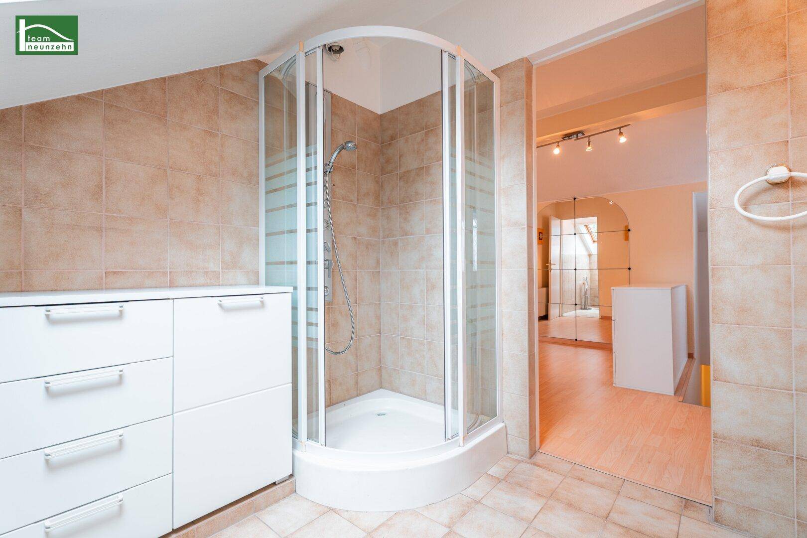 Helles Badezimmer mit Eckdusche, weißen Schränken und beigefarbenen Fliesen unter einer Dachschräge.