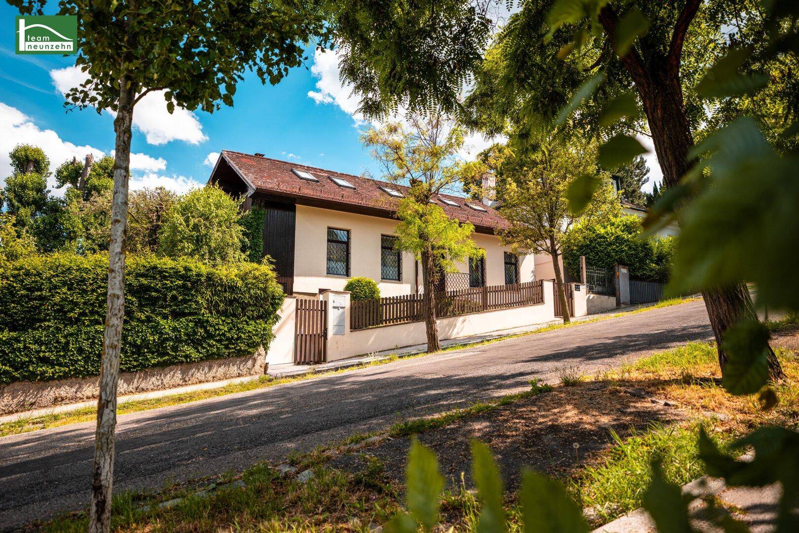 Charmantes Haus von der Straße aus gesehen, umgeben von üppigem Grün und Bäumen an einem sonnigen Tag.
