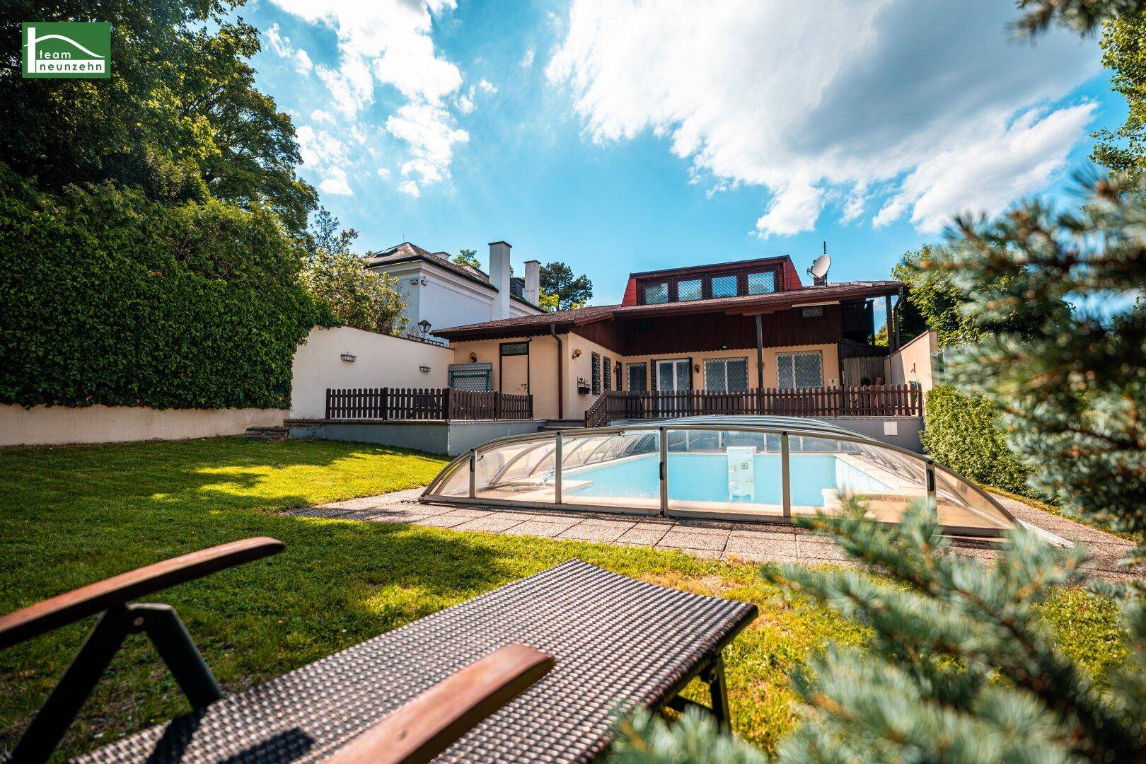 Großer Garten mit überdachtem Swimmingpool, Liegestühlen und dem Haus im Hintergrund an einem sonnigen Tag.