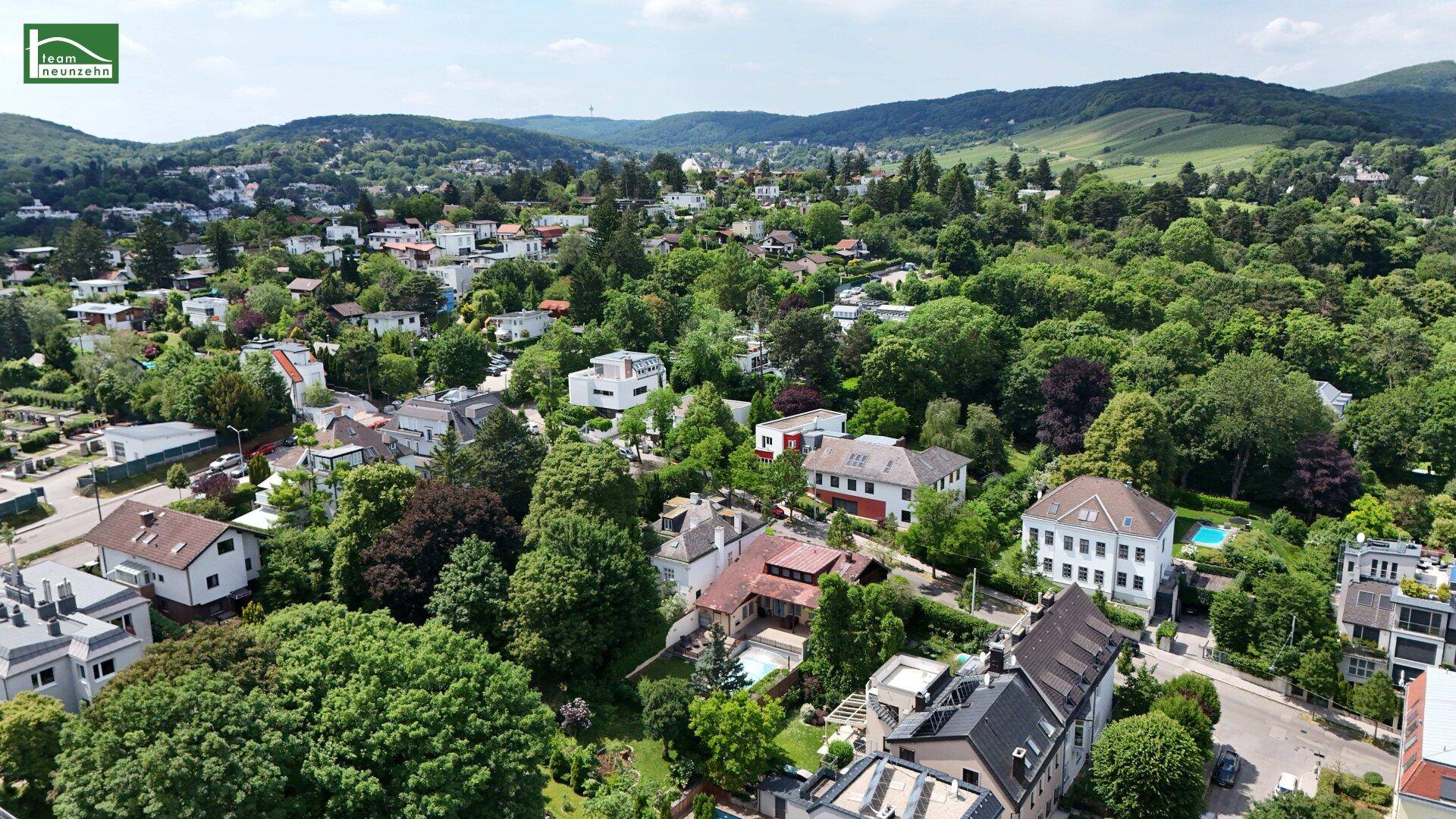Weitläufige Luftaufnahme einer grünen Wohngegend mit vielen Häusern und bewaldeten Hügeln im Hintergrund.