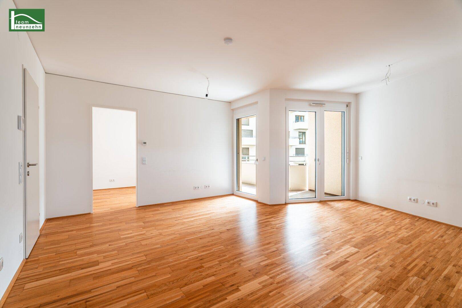 Helles, leeres Wohnzimmer mit Parkettboden und Zugang zu einem Balkon.