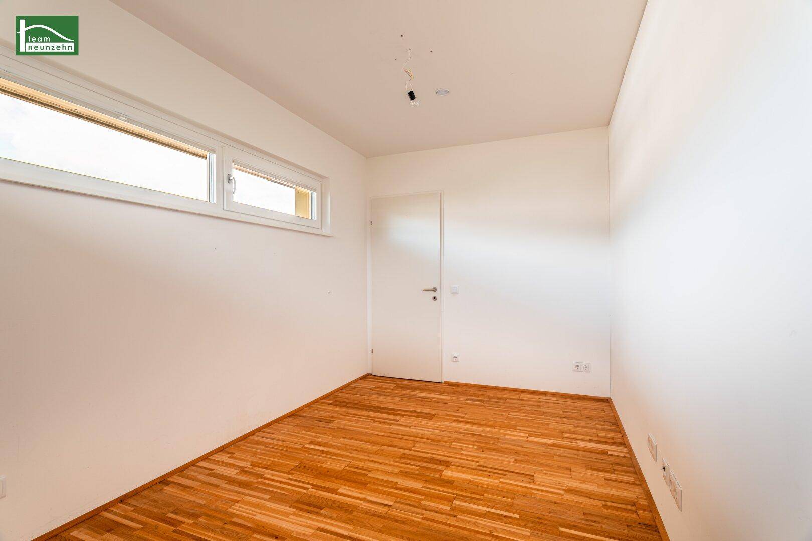 Leeres Zimmer mit Parkettboden und einem Fenster, das viel Tageslicht hereinlässt.