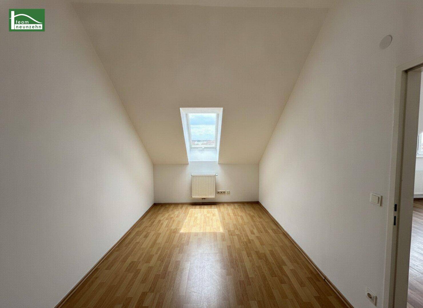 Langes, helles Zimmer im Dachgeschoss mit Laminatboden und einem Dachfenster.