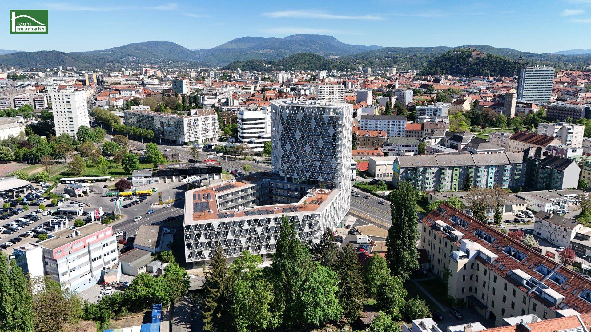 Panoramablick auf eine Stadt mit einem modernen Gebäude im Zentrum und Bergen im Hintergrund unter blauem Himmel.