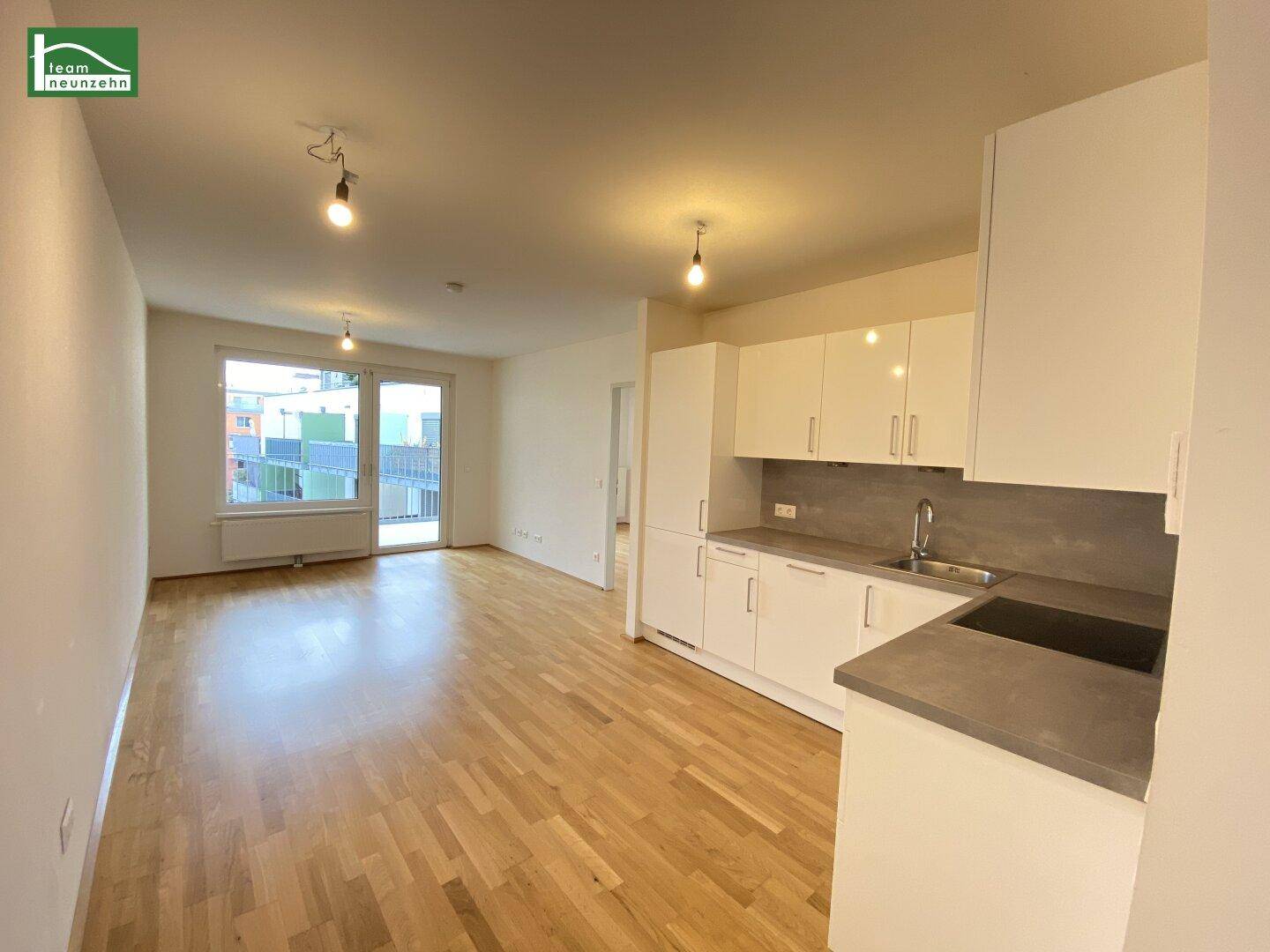 Heller offener Wohnbereich mit integrierter Küchenzeile, Parkettboden und Zugang zum Balkon.