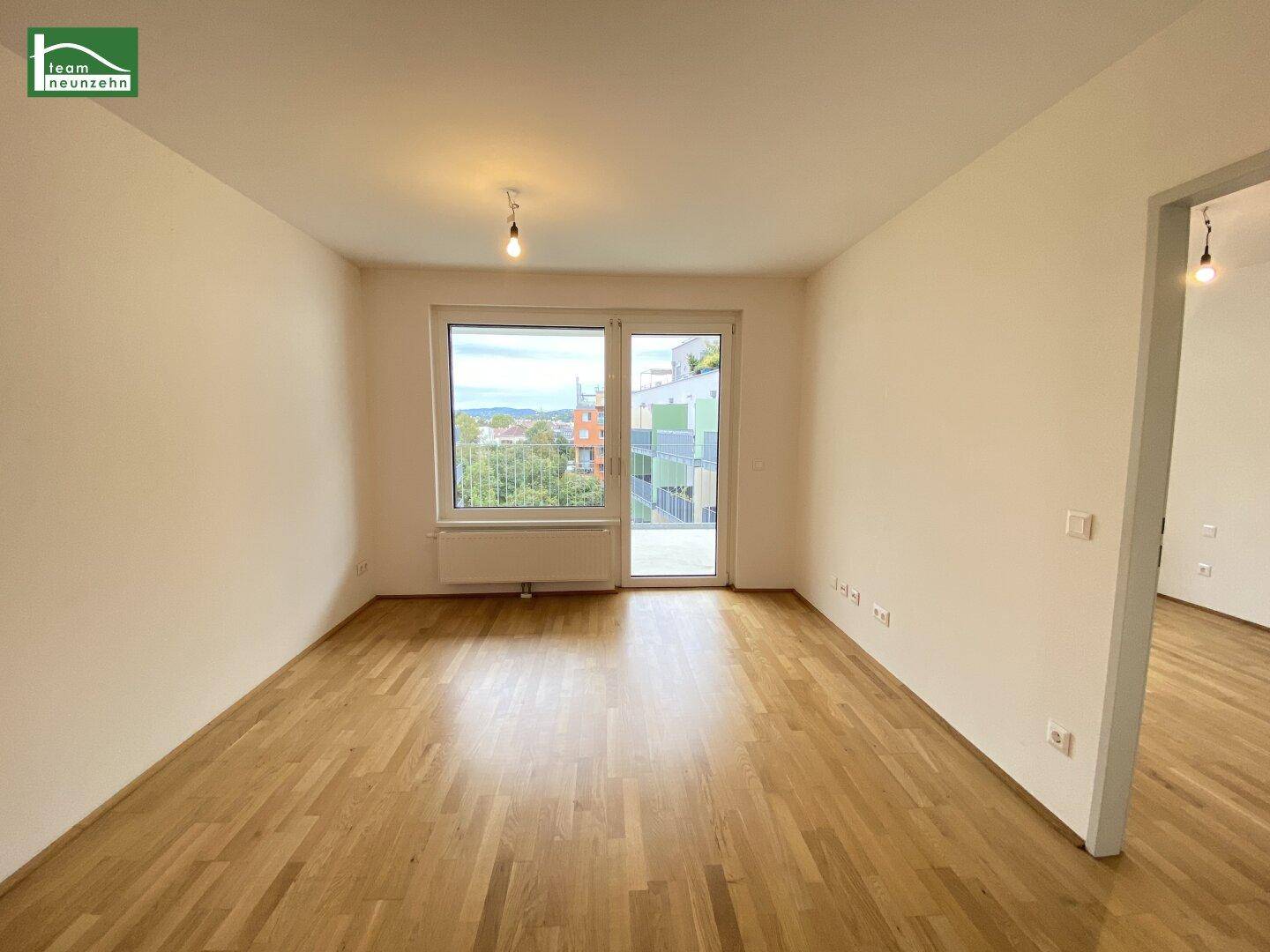 Leeres Zimmer mit hellem Parkettboden und großer Fensterfront, die viel Tageslicht hereinlässt.