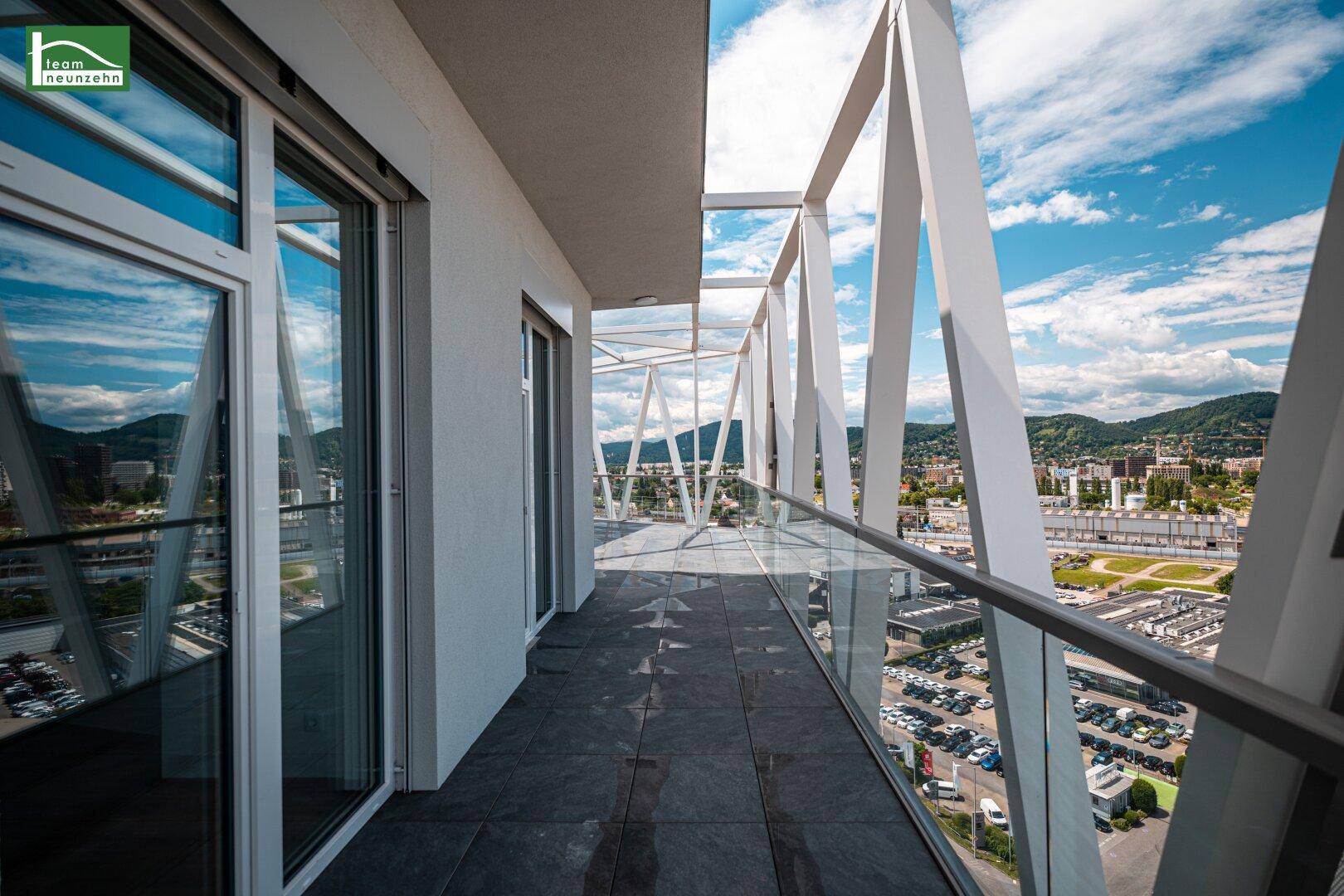 Langer Balkon mit Glasgeländer und Blick auf die Stadt und die umliegenden Berge.