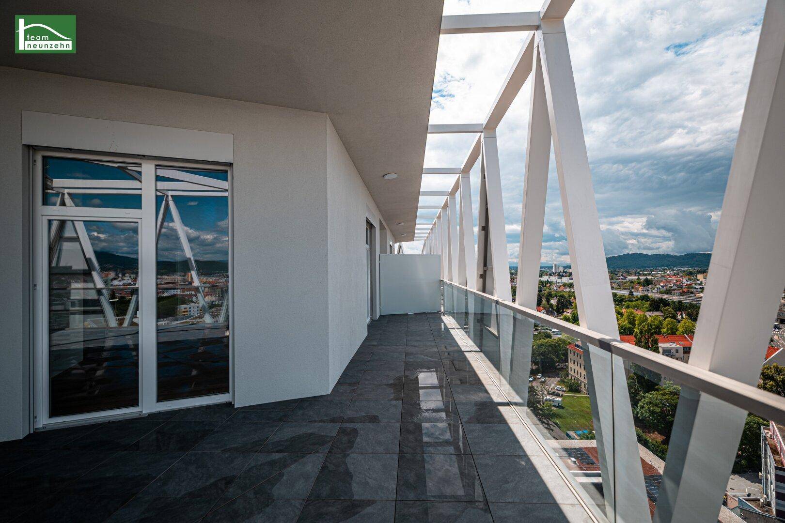 Langer Balkon mit Glasgeländer und Blick auf die Stadt und die umliegenden Berge.