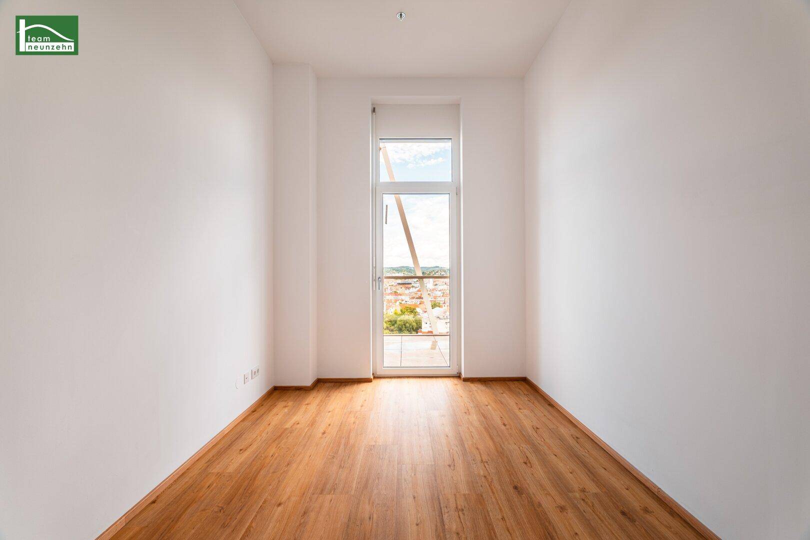 Heller Raum mit Holzboden und einer großen Glastür, die den Blick auf die Stadt freigibt.