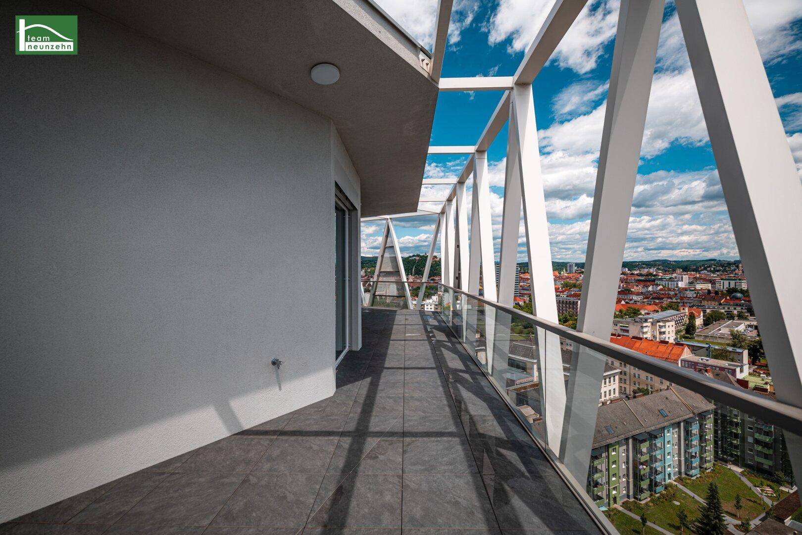 Langer, moderner Balkon mit Glasgeländer und freiem Blick auf die umliegende Stadtlandschaft.
