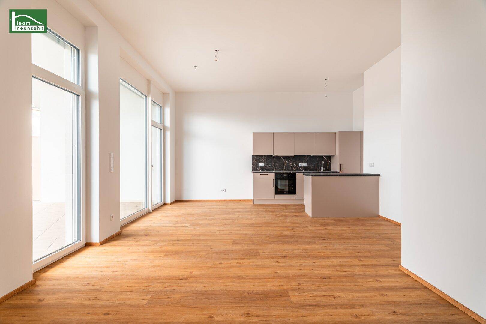 Heller offener Wohnbereich mit integrierter Küche, Holzboden und großen Fenstern zum Außenbereich.