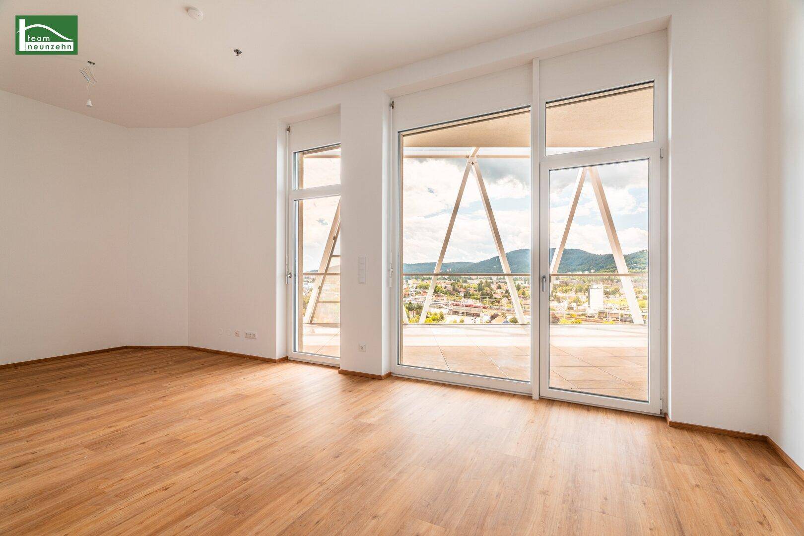 Geräumiges Wohnzimmer mit hellem Holzboden, großen Fenstern und Zugang zu einem Balkon mit Aussicht.