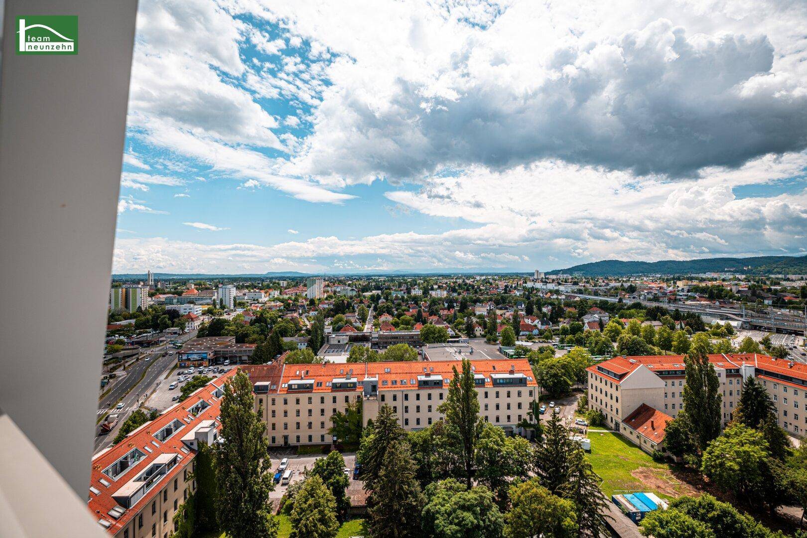 Weitläufiger Panoramablick über die Stadt mit grünen Bäumen und Bergen im Hintergrund unter blauem Himmel.