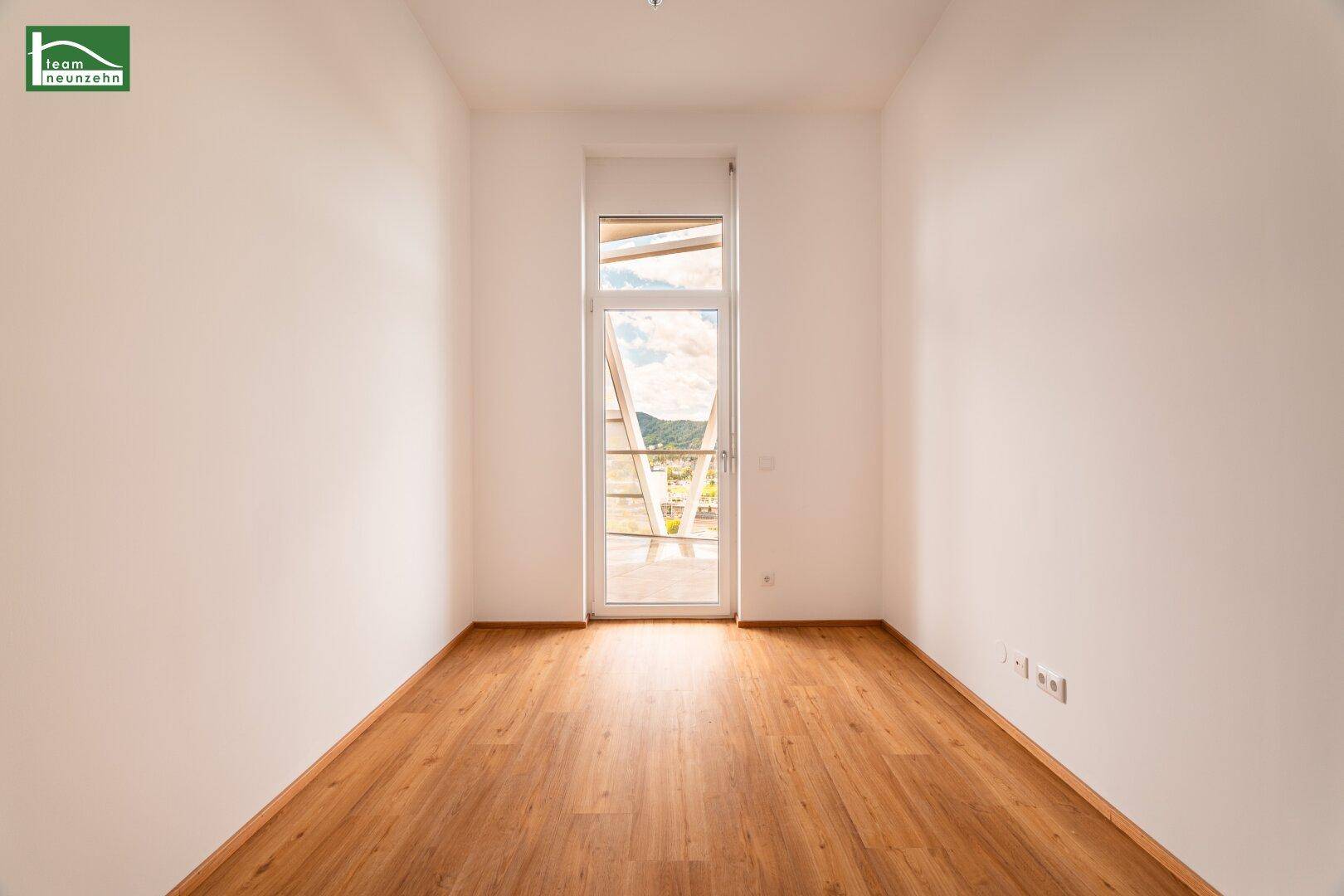 Heller Raum mit Holzboden und einem hohen Fenster, das viel Tageslicht hereinlässt und eine Aussicht bietet.