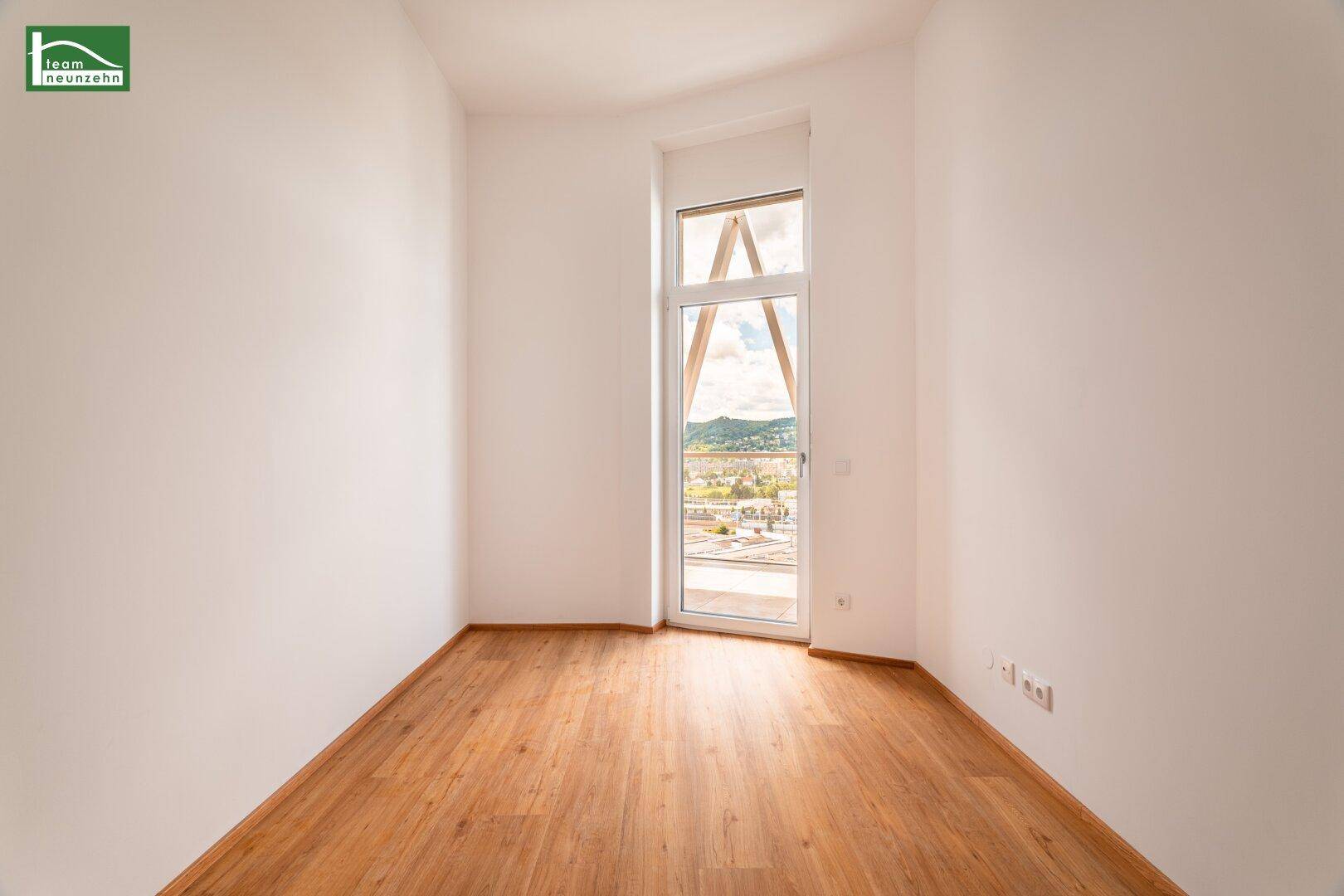Lichtdurchfluteter Raum mit Holzboden und einem hohen Fenster, das einen weiten Blick ermöglicht.