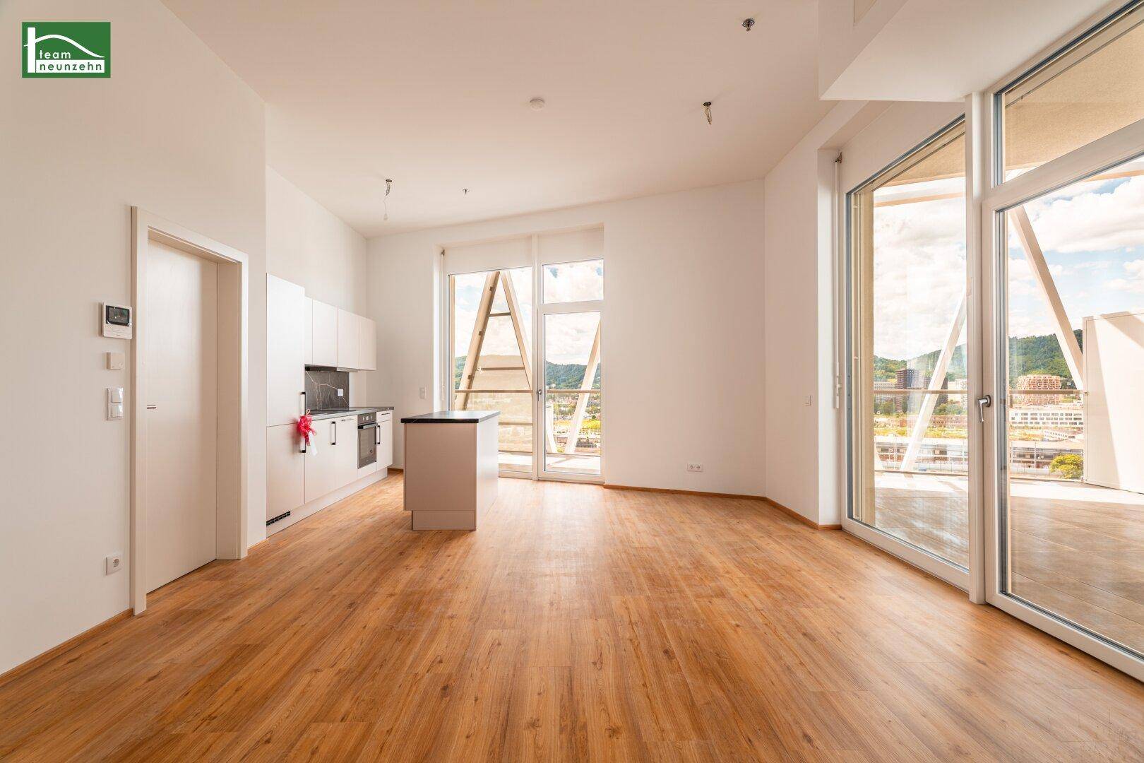 Offener Wohnbereich mit integrierter Küchenzeile, Holzboden und großen Fenstern mit Zugang zum Balkon.