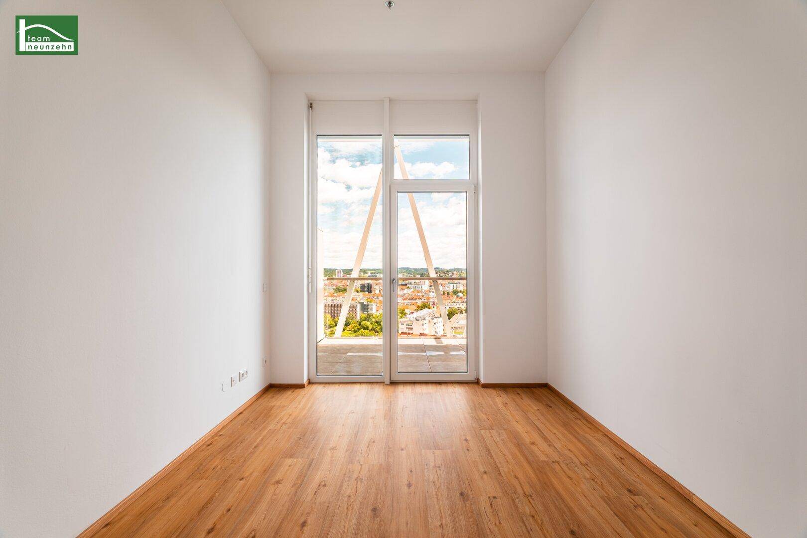 Heller Raum mit Holzboden und großem Fenster, das einen weiten Blick über die Stadt bietet.