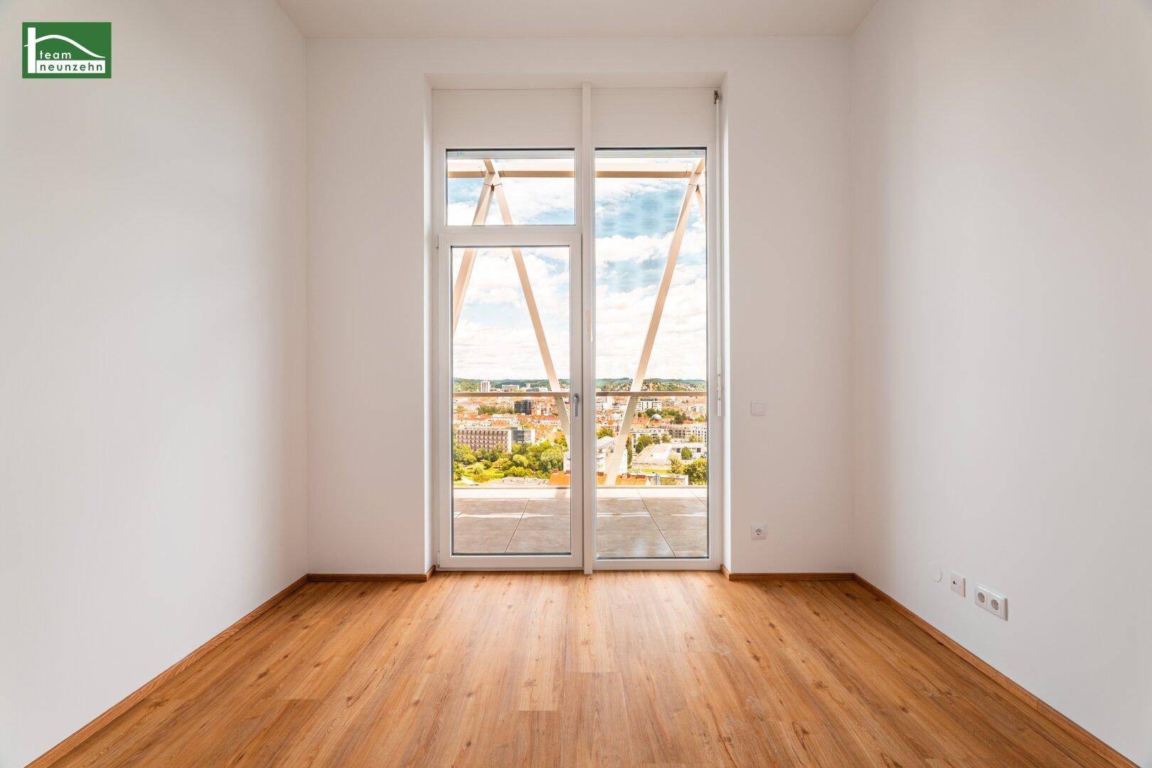 Geräumiger, heller Raum mit Holzboden und Zugang zu einem Balkon mit Stadtpanorama.