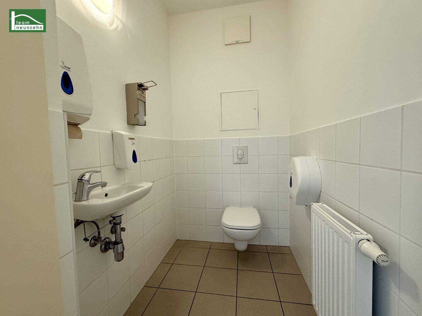 Separate Toilette mit Wand-WC, kleinem Waschbecken und weißen Fliesen an Wänden und Boden.