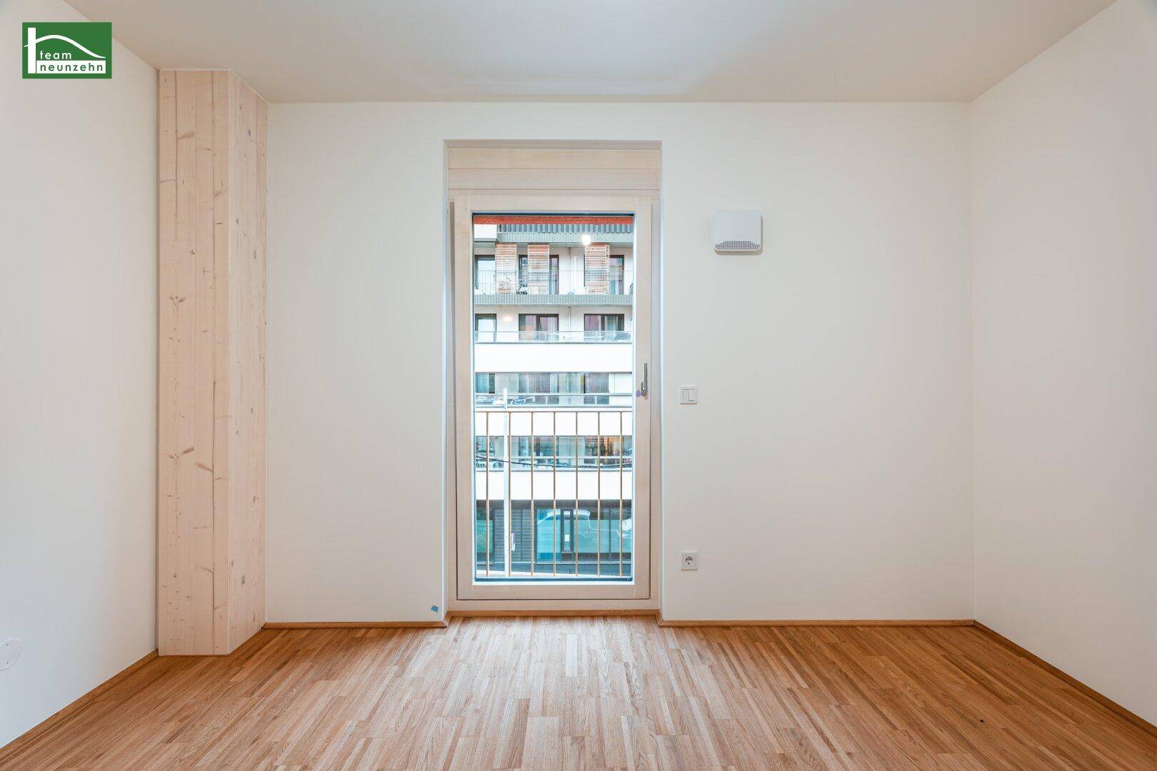 Helles Zimmer mit Holzboden und großem Fenster mit Blick auf das Gebäude gegenüber.