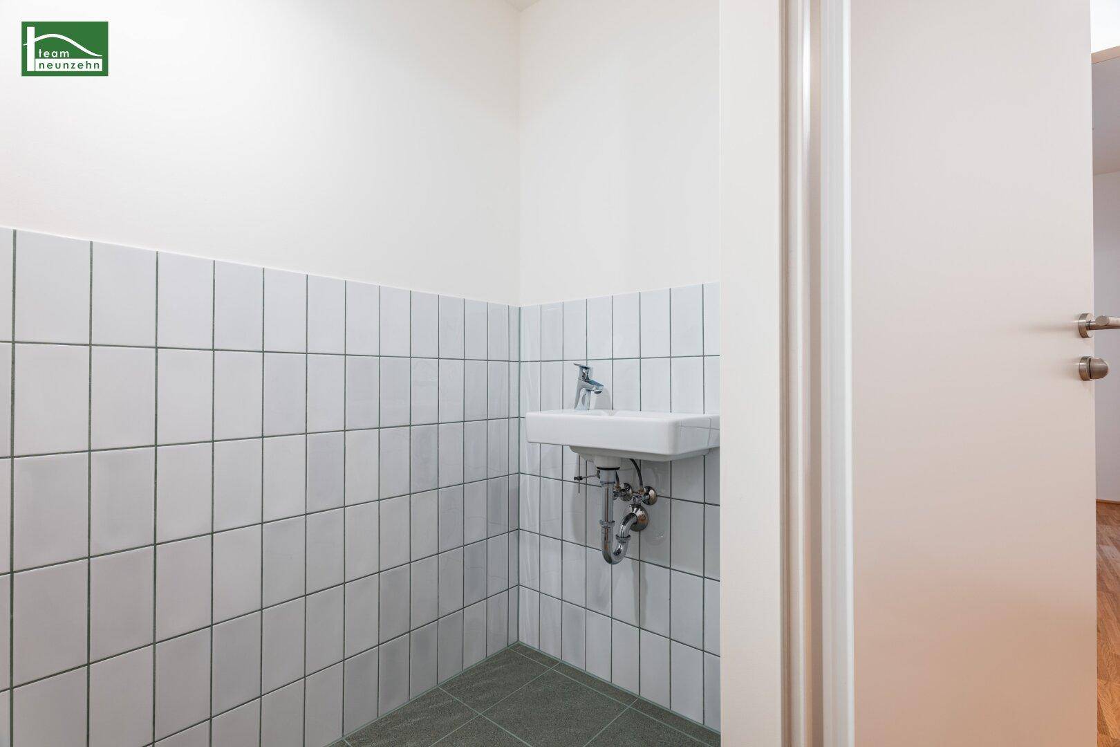 Kleines WC mit Waschbecken, weißen Fliesen an der Wand und grauem Fliesenboden.