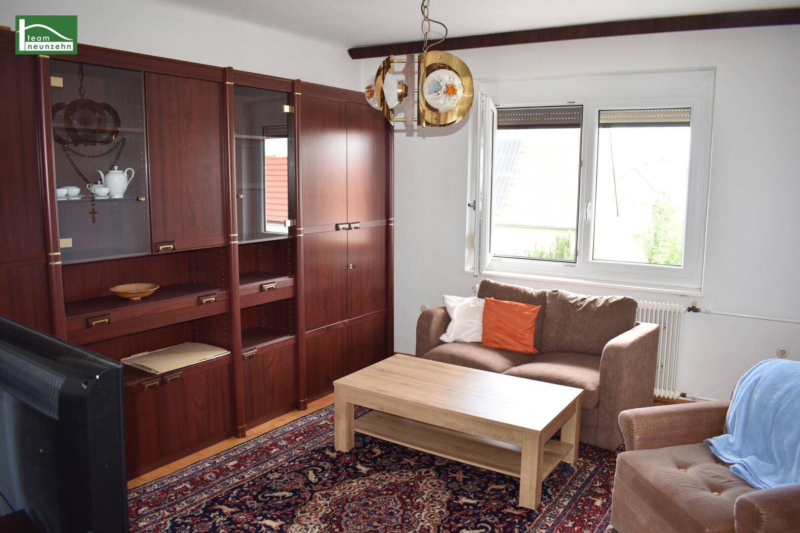 Gemütliches Wohnzimmer mit dunklen Holzmöbeln, Teppich und Fenster mit Blick ins Grüne.