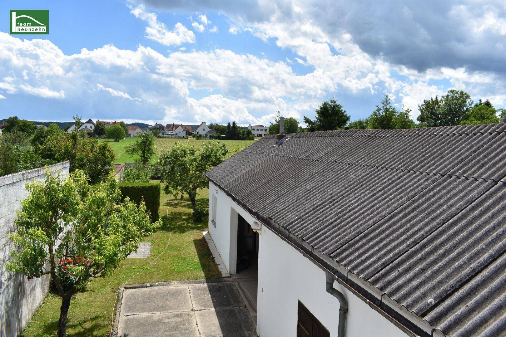 Blick von oben auf den Gartenbereich mit Rasen, Bäumen und einem Nebengebäude unter blauem Himmel.