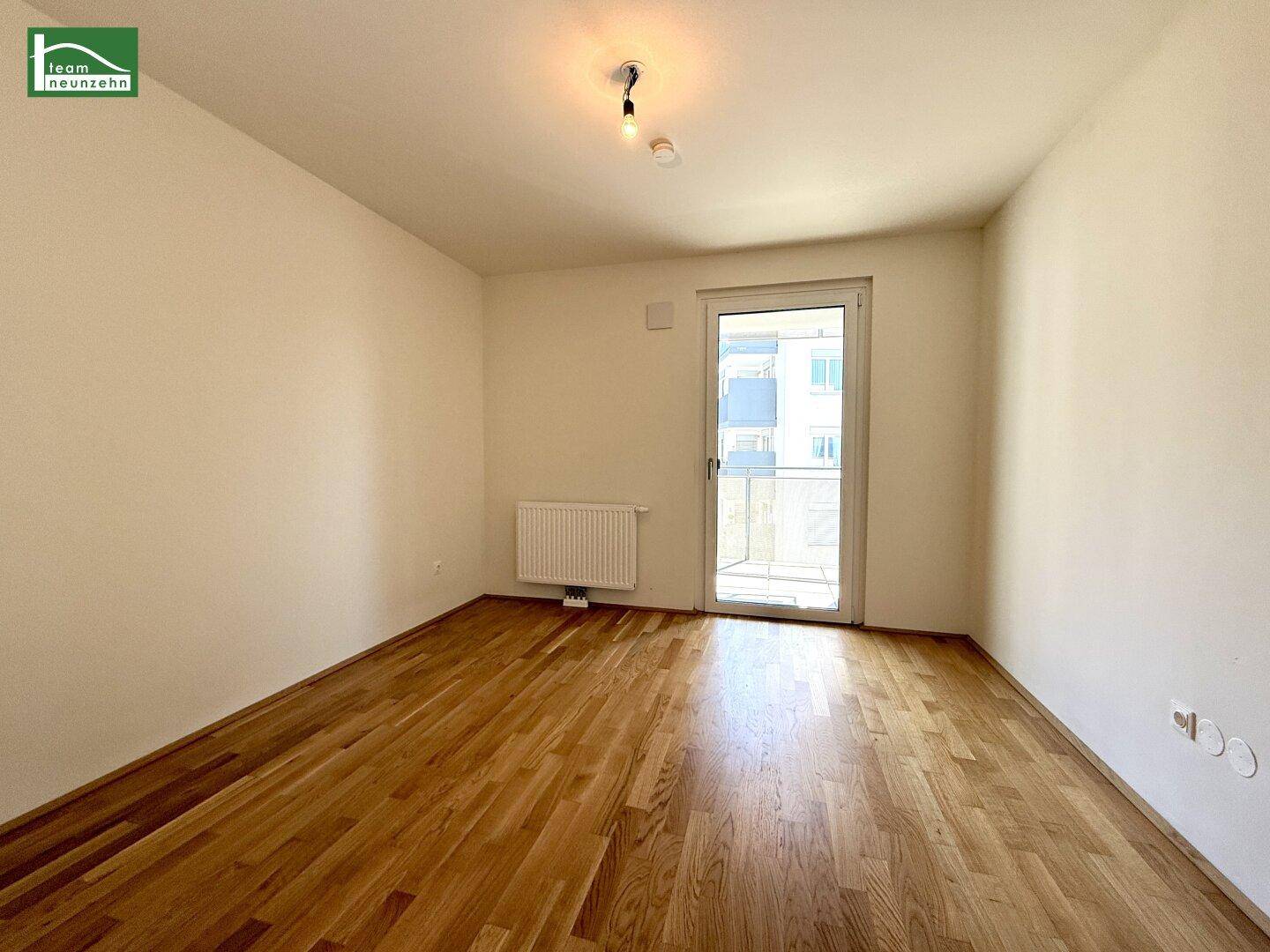 Leeres Zimmer mit Holzboden, Heizkörper und Zugang zu einem Balkon.