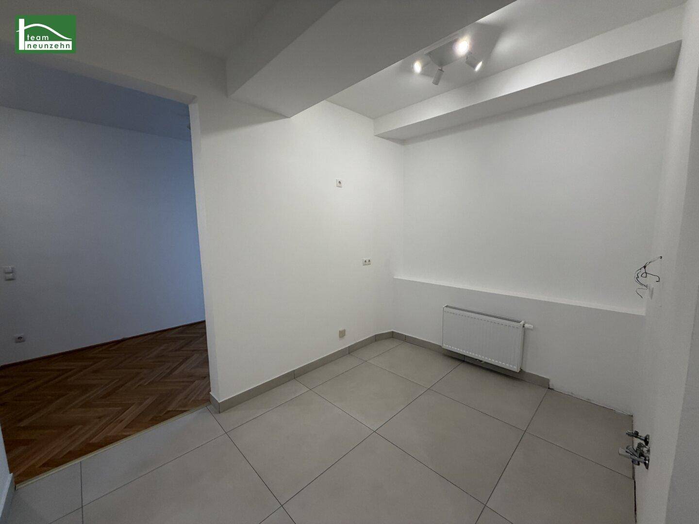 Leerer Raum mit hellgrauen Fliesen und einem Heizkörper, angrenzend an ein Zimmer mit Parkettboden.