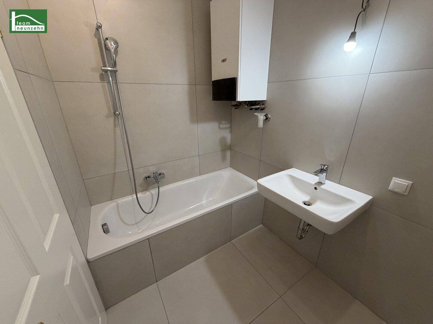 Modernes Badezimmer mit Badewanne, Duscharmatur und Waschbecken, komplett in hellgrauen Fliesen gehalten.