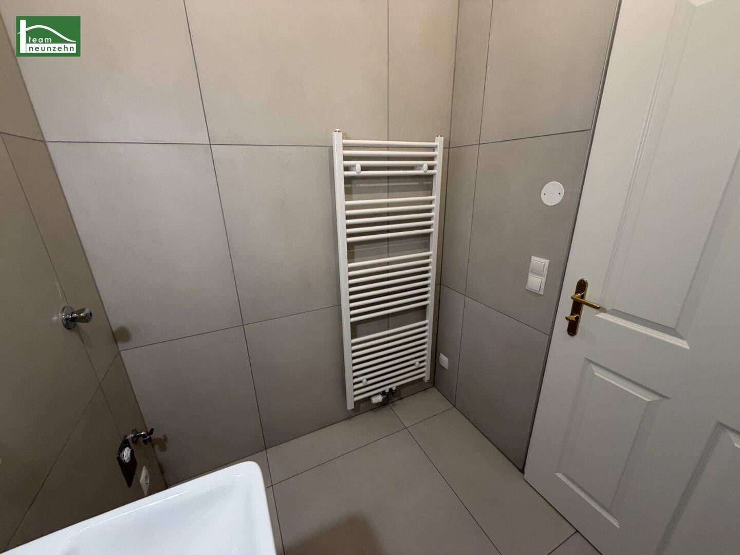 Badezimmer mit beheiztem Handtuchhalter und hellgrauen Wand- und Bodenfliesen, bietet modernen Komfort.