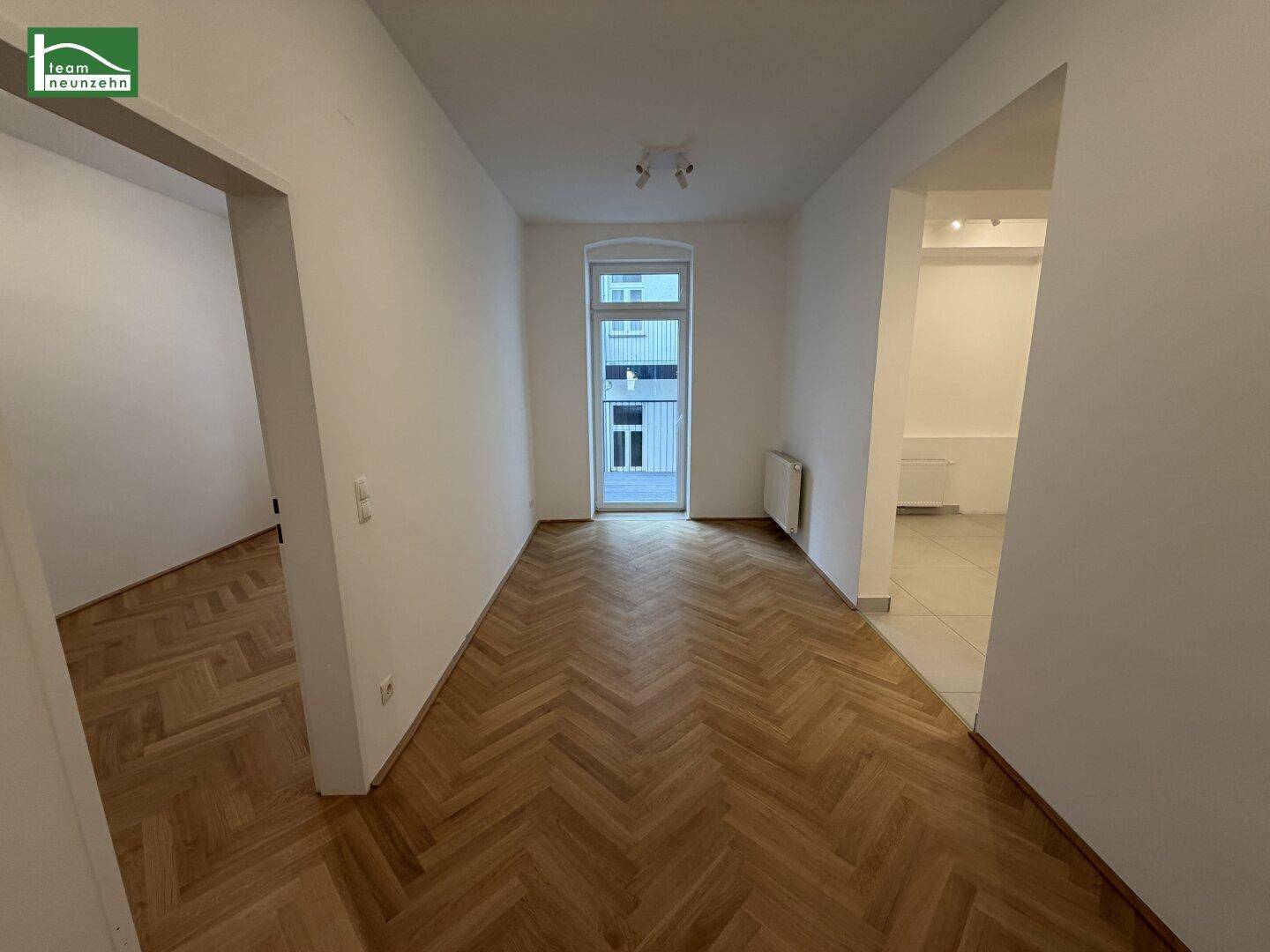 Geräumiger Innenraum mit elegantem Fischgrätparkett, großem Fenster und Zugang zu weiteren Zimmern.