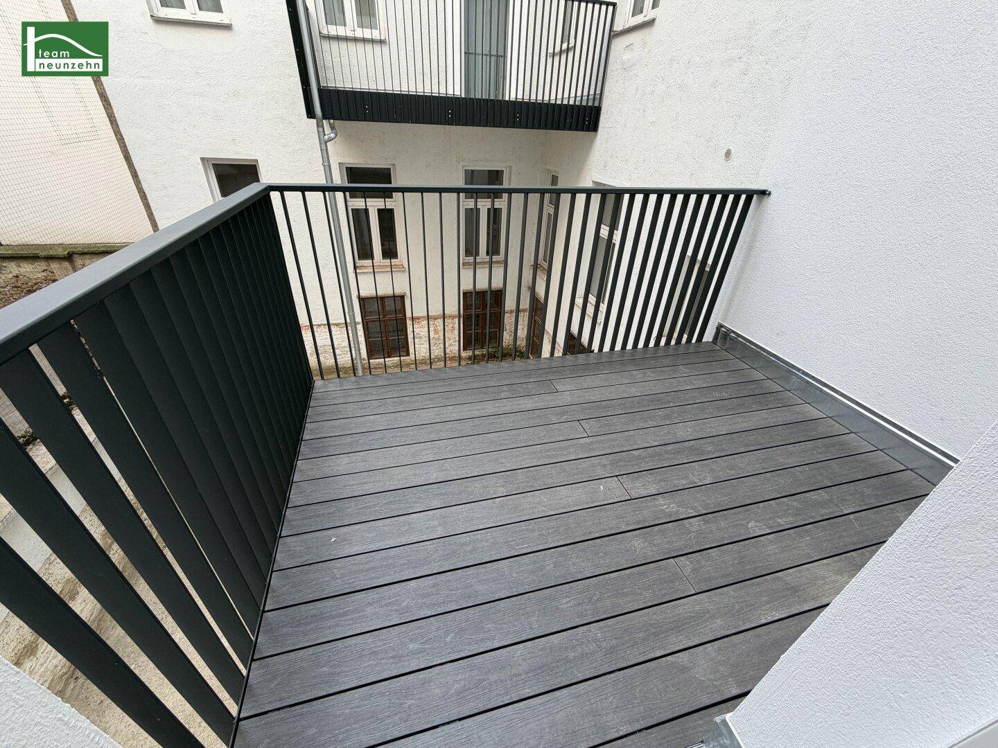 Moderner Balkon mit dunklem Bodenbelag und schwarzem Metallgeländer, umgeben von Gebäuden im Innenhof.