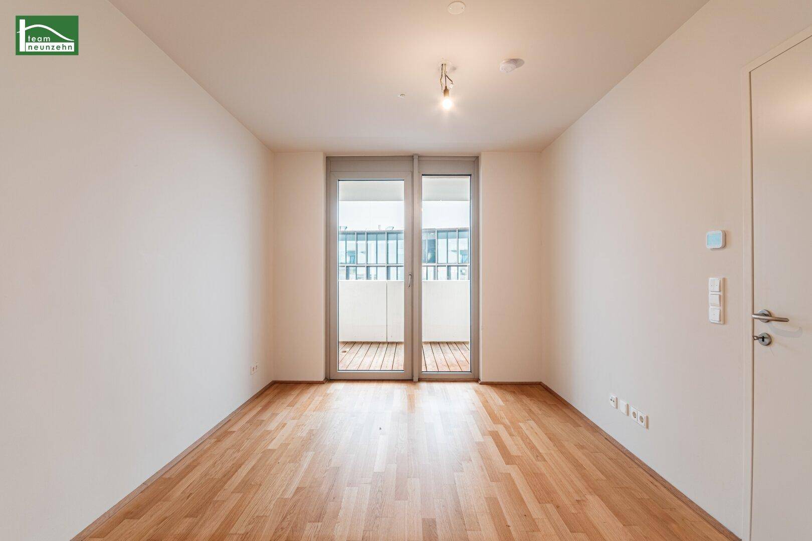 Helles Zimmer mit Holzboden und Zugang zu einem Balkon durch große Glasschiebetüren.