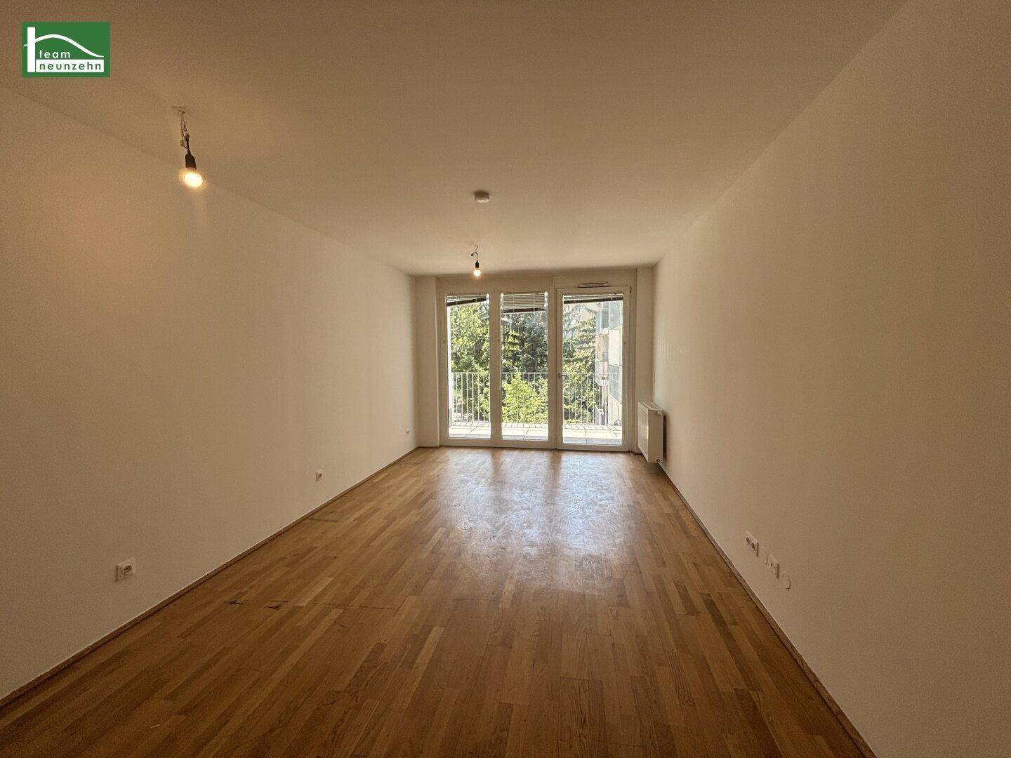 Geräumiges, unmöbliertes Wohnzimmer mit Parkettboden und großen Fenstern zum Balkon.
