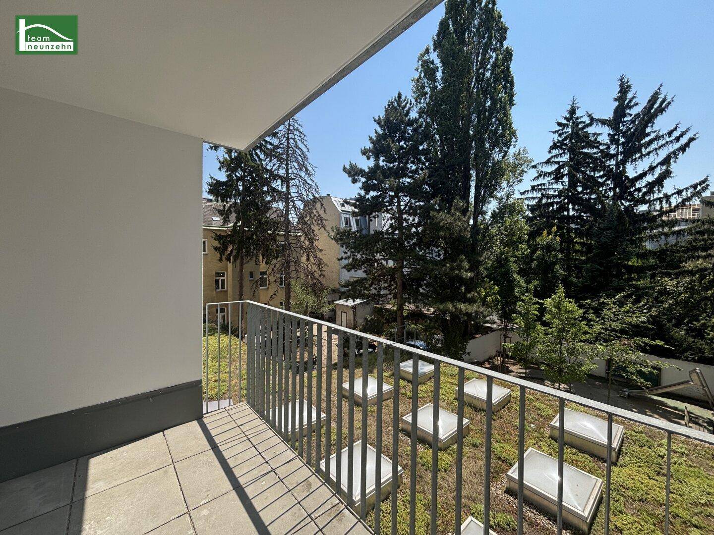 Sonniger Balkon mit Metallgeländer und Blick auf eine begrünte Dachlandschaft und Bäume.