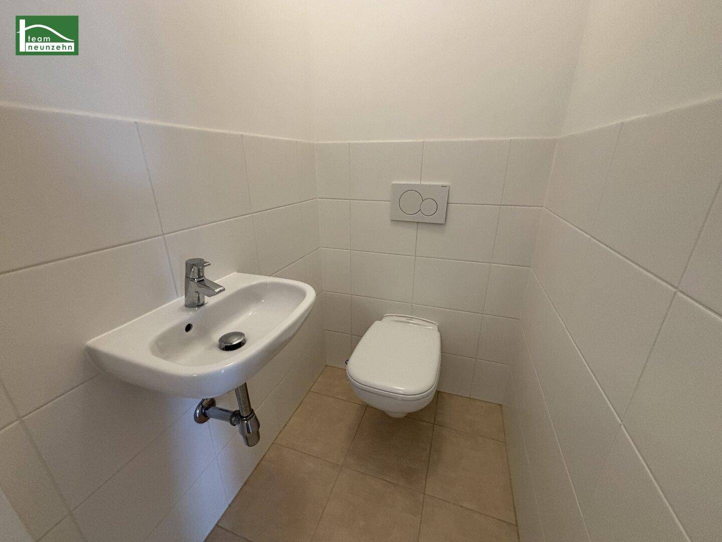Separates WC mit weißen Fliesen, wandhängender Toilette und kleinem Handwaschbecken.