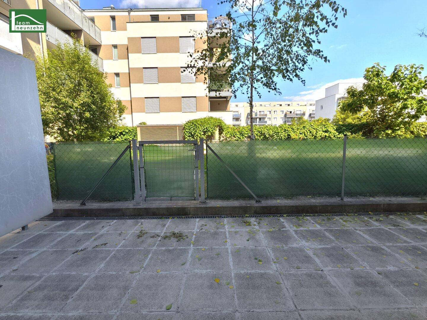 Gepflasterte Terrasse mit grünem Zaun und Blick auf die umliegenden Wohngebäude.