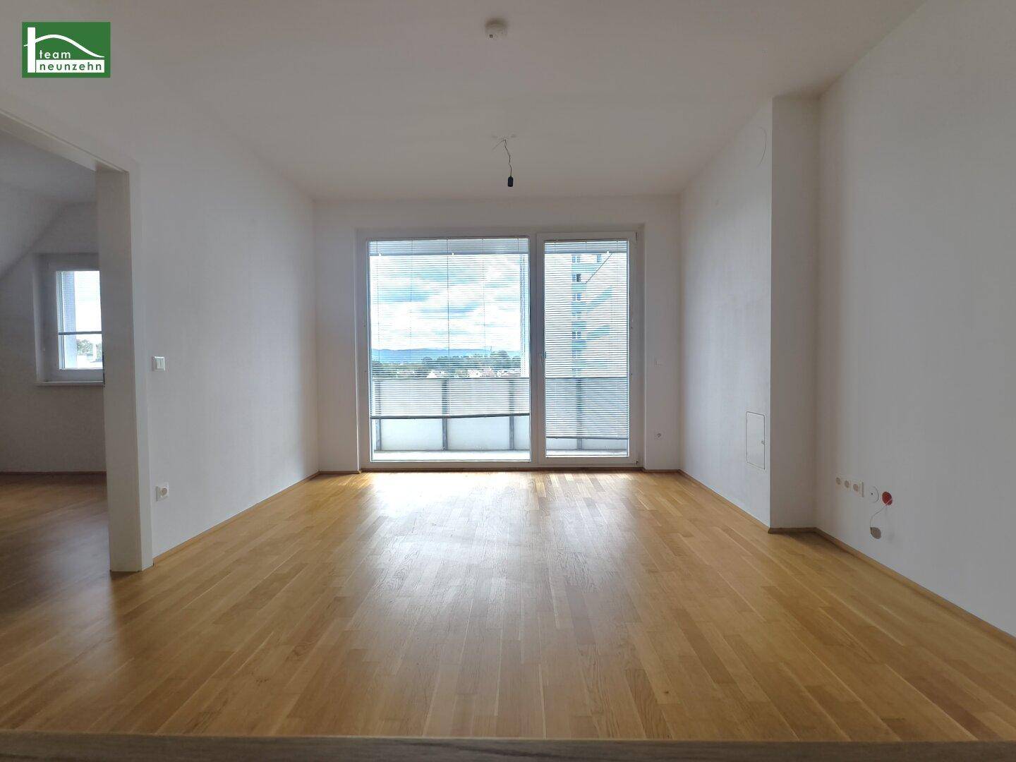 Heller Wohnraum mit großen Fenstern, Holzboden und Zugang zu einem weiteren Zimmer.